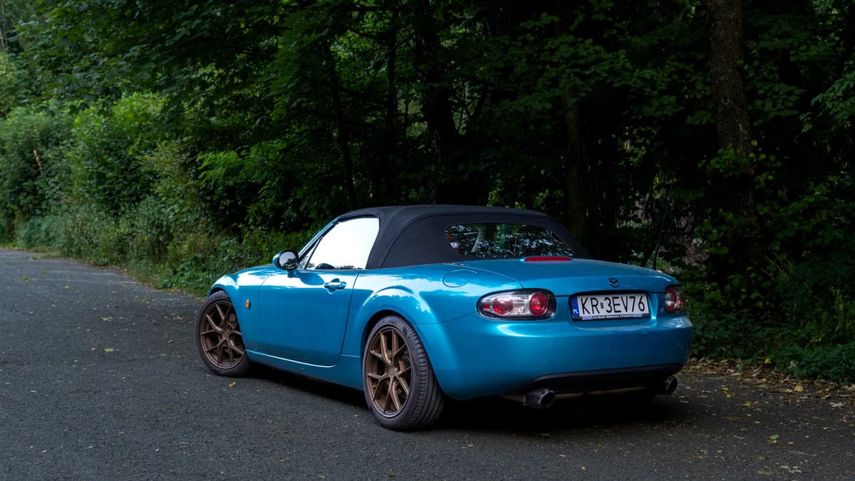 Blue Mazda MX-5 convertible parked on a forest road, showcasing its sporty design.